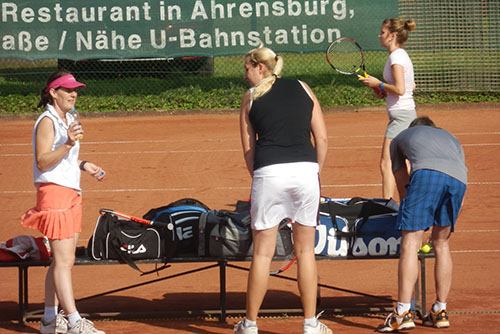 Drei Frauen stehen auf einem Tennisplatz, vor ihnen eine Bank mit Tennisausrüstung. Hinter der Bank befindet sich eine weitere Frau, die bereits spielt.