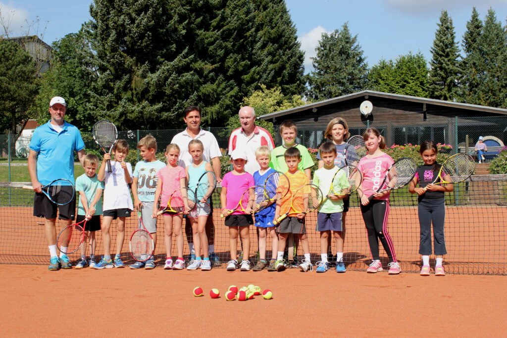 Eine Gruppe Kinder steht vor einem Tennisnetz; hinter ihnen stehen drei Tennistrainer.