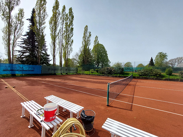 Ein Tennisplatz mit weißen Bänken im Vordergrund. Rechts auf dem Bild ist ein Tennisnetz zu sehen, das über das Spielfeld gespannt ist.